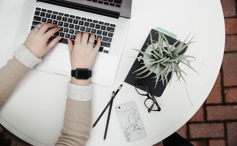 Typing the ideal query letter length at a laptop with plant, glasses, phone, pencils