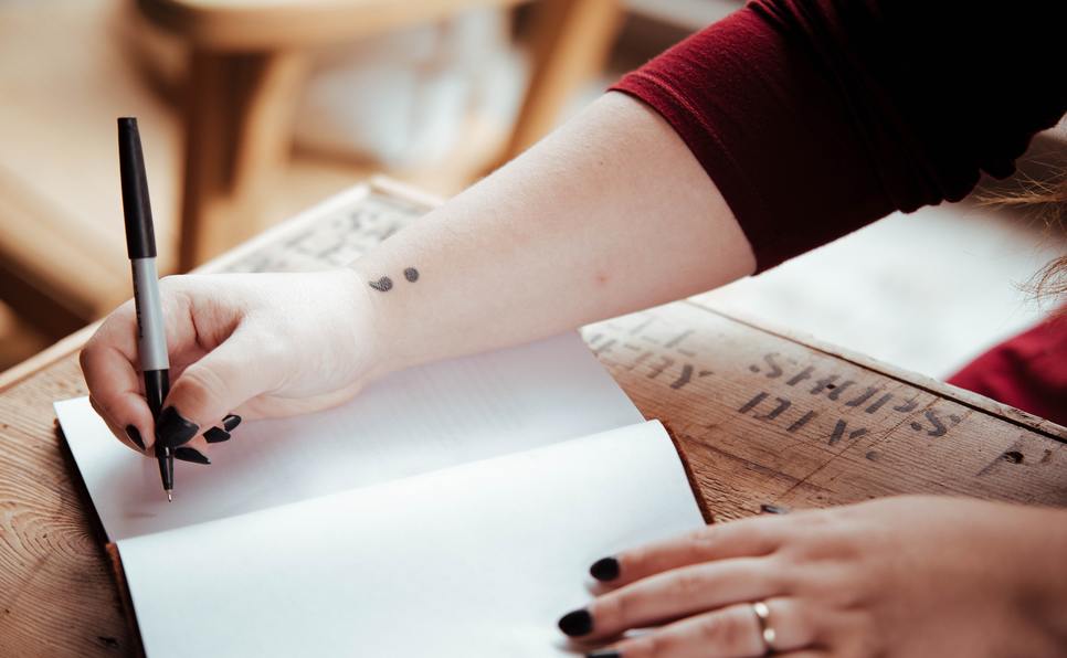 A woman's wrist with a semi-colon tattoo writing in a blank book representing how to choose a literary agent for your entire writing career